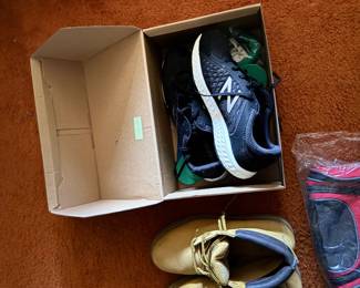 Pair of black New Balance sneakers in an open cardboard box alongside tan work boots resting on shag carpet.