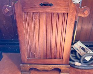 Vintage wooden tilt-out trash bin or hamper featuring beadboard paneling, decorative side handles, and a black metal pull, shown with decor items stored inside.