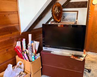 Vizio flat-screen LCD television resting on a low wooden chest, accompanied by cardboard boxes filled with wrapping paper rolls under an angled ceiling.