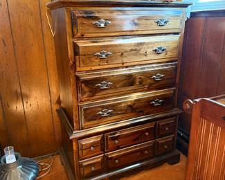Vintage dark-stained pine chest-on-chest tall dresser with brass batwing hardware, topped with decorative dolls and an oil lamp.