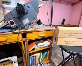 Workshop area featuring a grey tabletop power tool, a Dremel Drill Press attachment box, a cord reel with yellow extension cord, and various wooden desks and storage cases.