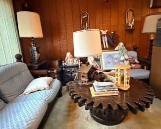 Vintage wood gear-shaped coffee table surrounded by a blue upholstered sofa, armchair, and table lamps in a paneled living room setting.