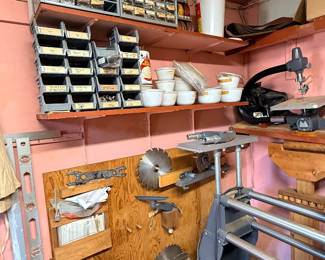 Workshop storage setup featuring metal drawer organizers, stacked part bins, table saw blades mounted on wall panels, and stationary woodworking machinery.