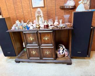 Traditional dark wood media console featuring a central cabinet, brass hardware, flanking Sharp floor speakers, and a tabletop displaying assorted porcelain figurines and glass decor.
