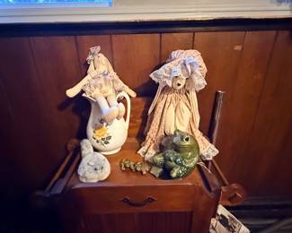 Vintage soft-sculpture rabbit dolls resting on a ceramic pitcher and wooden nightstand, alongside ceramic frog figurines and a plush cat.