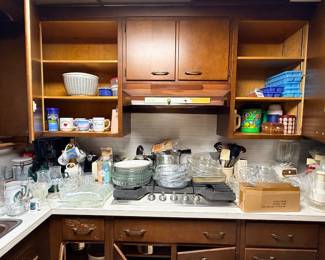 Assorted vintage kitchenware, including glassware, mugs, cookware, and small appliances, displayed on counters and open wooden cabinetry.