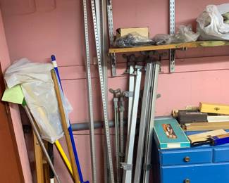Assortment of long aluminum bar clamps leaned against a wall in a workshop area alongside cleaning tools and a blue chest of drawers.