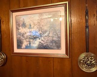 Framed landscape art print of a forest stream, flanked by a pair of vintage decorative brass warming pans with wooden handles.