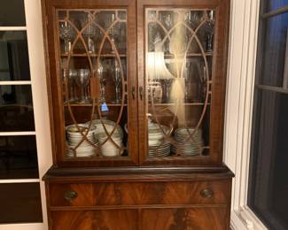 American Chippendale revival china cabinet from the 1930s or 1940s. Broken swan-neck pediment and flame mahogany veneer on lower doors; oval brass pulls and decorative glazing.
