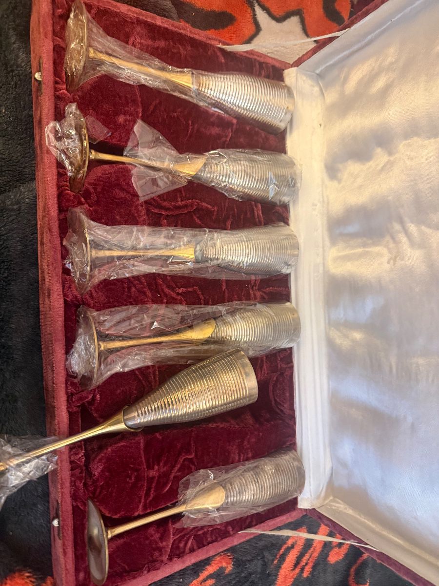 A stunning vintage hospitality set featuring 6 metal champagne flutes. Each glass has a beautifully textured, ribbed silver-tone body paired with a sleek gold-tone stem and base. The set comes protected in its original, luxurious deep red velvet presentation display box with a clean satin-lined interior lid. Most glasses are still individually wrapped in plastic