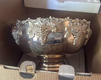 • centerpiece bowl from the silver-plated beverage cooler set. This photo highlights the beautiful, deeply embossed grape clusters and vine leaves that line the scalloped rim. It shows the bowl securely nestled in its original cardboard and styrofoam protective packing box, proving it has been carefully stored. Perfect for buyers who want a clear look at the intricate metal detailing and reflective finish.