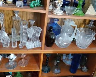 A spectacular multi-shelf collection of vintage glassware, perfect for dealers or collectors. Filled with dozens of premium pieces including matched sets of cut-crystal glass candlesticks, formal serving decanters, decorative colored glass taper holders (in deep blues and vibrant greens), a ceramic multi-candle candelabra, and various artistic glass vases.