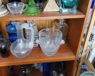 Beautiful selection of fine glassware and vintage barware. Features a clear cut-crystal ice bucket with side handles, elegant glass spirits decanters with stoppers (including a unique spiral-handled clear glass pitcher), deep cobalt blue glass candle holders, an interesting frosted green depression-style glass pedestal dish, and a teal blue square glass decanter bottle.