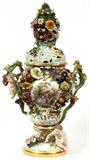 Lot#1003, MEISSEN PORCELAIN COVERED URN, C. 1900, H 26", W 14"A hand painted courting scene at one side and a floral spray on the reverse, flanked by stem handles with applied putti and flowers, the cover centered by a large floral finial, marked in blue under glaze with crossed swords (see additional photo).