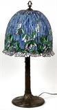 Lot#1017, TIFFANY STYLE LEADED GLASS LAMP, H 36" W 14"Raised on a trunk form base, with leaded glass shade decorated in purple floral motif.