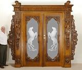 Lot 1051:  A GRAND CARVED AND ETCHED FRENCH DOORS ENTRYWAY: Heavily carved all over to include 4 urn finials over lentil with Northwind masquerons. Frame with figural bust, foliate decoration. Removable side panels with foliate and putti figures. Each door with carved bezel, mirror image etched glass dancing horses. Overall approx. 116'' h. x 116'' wide, each door 91 1/2'' h. x 38''.
CONDITION: Split in top lintel. All measurements are approximate, Burchard Galleries strongly advises to have your own contractor re-measure to make sure it will fit in your desired location.