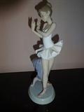 A Lladro ballerina depicted standing next to a stool. It is hand painted porcelain and stands 10" high by 4.5" long by 3" wide.