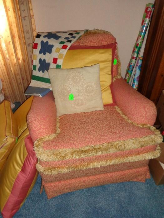 Pink print upholstered chair with crème fringe trim; yellow and pink throw pillows, several vintage quilts and vintage quilt top