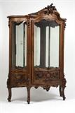 Lot# 1028 A Louis XV style carved walnut vitrine Late 19th / early 20th century, of serpentine outline, the arched, scrolled cornice centering a rocaille clasp, over a glazed and paneled door and side panels divided by flower and shell-headed uprights, enclosing a mirrored interior with glass shelves, raised on foliate scroll cabriole legs, electrified, with key, 82'' H x 59.25'' W x 26'' D, est: $2000/3000
