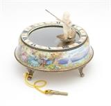 Lot# 1033 A Juvenia silver, gilt metal & enamel mystery clock Circa 1920, serial number ''58058'', hallmarked for .935 silver and marked ''Argent Dore'', signed: ''Ateliers Juvenia / a La Chaux-De-Fonds (Suisse) / et a Paris / fab. Suisse, Metal Dore, Metaux Divers'', further signed ''Juvenia Mfg. Swiss'' and ''DG'', the circular rotating mirrored platform supporting a carved putto standing on a silvered leaf and a holding a spear-shaped pointer to indicate time, surrounded by a circular dial with blue Roman numeral hour markers and pink rose blossoms, on an enameled silver circular base depicting Commedia dell' Arte figures dancing in a landscape, raised on foliate scroll feet, winds at bottom base, with key, 2.5'' H x 5.5'' Dia., est: $4000/6000