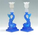 Lot 1088:  PAIR PRESSED GLASS DOUBLE STEP DOLPHIN CANDLESTICKS: Pressed glass with white single light holder over blue figural dolphin, stepped square base. Overall measures 10 3/4'' h. x 3 3/4'' square.
CONDITION: One with internal crack at top candle holder.