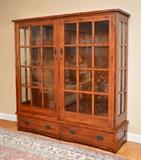 Lot 1123:  ARTS & CRAFTS MISSION STYLE DOUBLE DOOR OAK DISPLAY CABINET: 2 part cabinet, with doors having 12 panes of glass each. Illuminated Interior with glass shelves. Unusual touch light feature attached to outer hinge. Rests on the base with two drawers. Key locking. Later 20th C. Measures 68'' h. x 63'' x 18 1/2''.