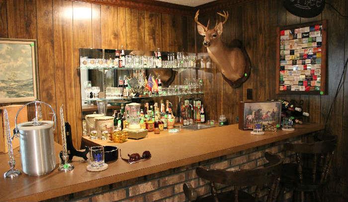 Close-up view of the bar in den with all glassware, wine crates, look at the wall hanging with a collection of matches. 