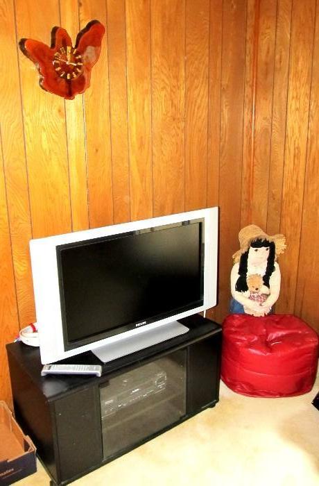 TV Stand with dark finish, glass door shelves storage.  Also the Emerson VHS Player is available; Phillips Flat Screen Color TV;  Also shown is a Small Poof with Red Leather like upholstery and a Craft Country Lass Doll with a stuffed bear are also available; The Cypress Clock is also available.