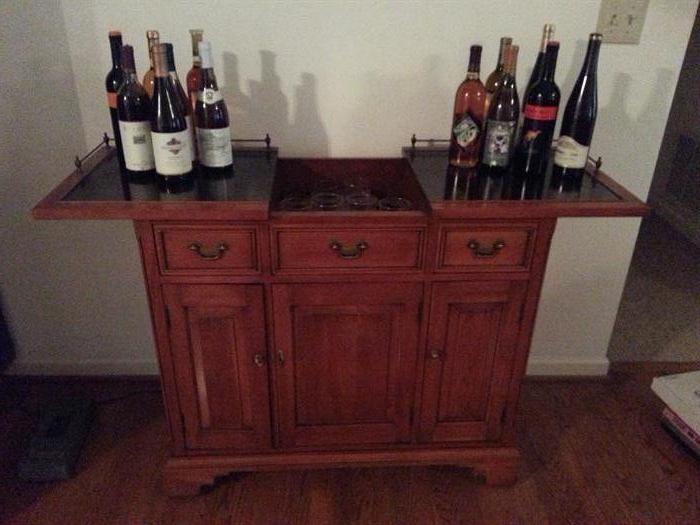 Bar with two drawers, two cabinet doors open to storage below and the top slides out to reveal storage of bar glassware.
12 bottles of local wine shown on bar.
