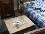 Small wood table with checker board on top. Blue plaid sleeper love seat and a goose magazine rack.