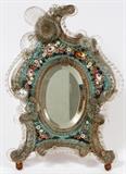 Lot#13, VENETIAN MICROMOSAIC EASEL BACK DRESSER MIRROR, H 16" W 12 1/2"Oval form mirror, measuring H. 6 1/4" x 4 1/2", framed in micromosaic clustered floral motif on a blue ground, and fitted with scrolling and floral form colorless glass ornamentation along the edge. Measuring H. 16" x 12 1/2" overall.