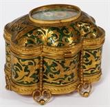 Lot#31, TAHAN FRENCH ENAMEL & GILT METAL BOX, LATE 19TH C., H 4", L 4 3/8"Footed lobed form dresser box, decorated in scrolling foliate motif and green enameled detailing. Featuring a hand painted floral bouquet plaque at the center, hinged lid opening to a ivory silk lined interior. Measuring H. 4" x 4 3/8" x 3 1/4". Original Tahan label at the underside.