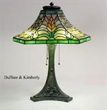 Lot 1051: DUFFNER & KIMBERLY 6 SIDED LEADED GLASS TABLE LAMP: Unique 6 sided leaded stained glass lamp with alternating panels of stylized wheat and geometric motifs on shade. Shade measures 6 1/2'' x 14 1/4''. Heavy cast bronze base with matching wheat pattern, 3 socket fixture, 21'' h. x 9''.
CONDITION: Several panels with fractures, dings and bends to heat cap.