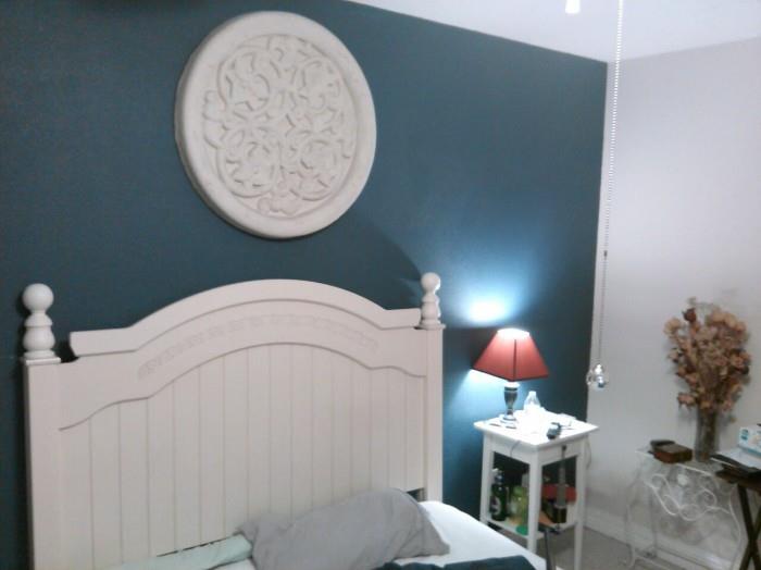 Wall medallion, cream headboard, lamp, 1 of 2 white side tables, white metal stand, queen bed partial shown