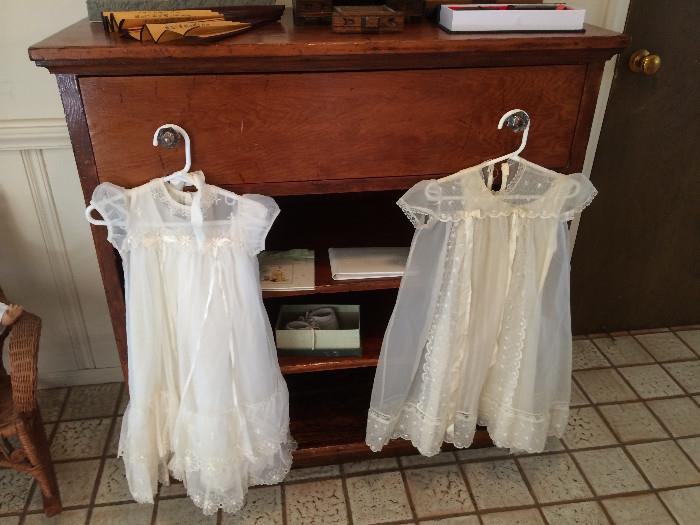 2 White Christening Dresses hanging from an Oak Dresser. Baby Brag Books. White Baby Shoes.