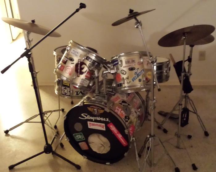 Slingerland Drum Set, newer, not-vintage: 5-pc with Slinglerland-labeled high-hat & cymbals, includes extra Tama Bass Pedal, Pearl COW BELL, an old Vintage Jazz Crash Cymbal and sticks/stick-bag (Silver, but covered in Stickers representing 80’s & 90’s counter-culture bands / skateboards / BMX).