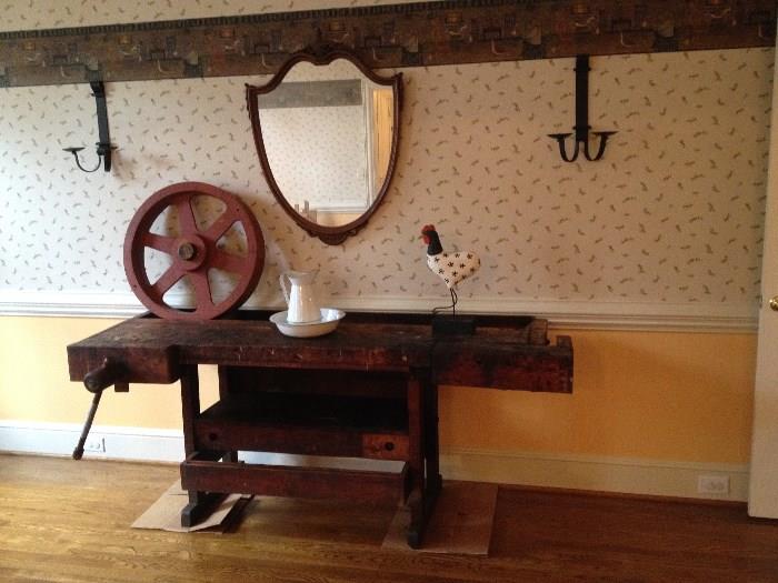 Antique work bench with vise holding old red industrial wheel, enamel pitcher and bowl, a carved rooster, shield shaped mirror and a pair of hanging hammered iron sconces