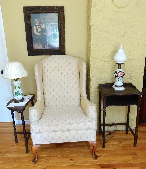 Wingback chair with Queen Anne wooden legs.  Chair is staged beside an antique half moon accent table complimenting white glass leaf design lamp. Telephone table with vintage white globe lamp rose painted design sits on vintage telephone table.  
