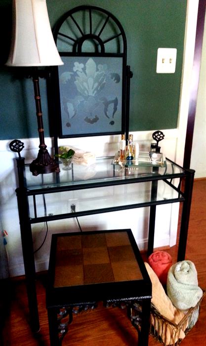 Black metal vanity with stool, contemporary lamp, perfume bottles, ladies watch, towels, wire basket.