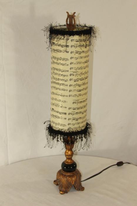 Furnishing – Table top lamp. Piece has long shade which is a piece of sheet music. Black fringes on top and bottom. Small lamp base. Metal with black embellishment. Nice piece