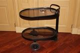 Furnishing – Small oval end table on wheels. Piece has a black metal frame with glass shelves. Piece is two tiered