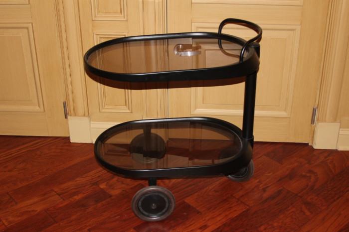 Furnishing – Small oval end table on wheels. Piece has a black metal frame with glass shelves. Piece is two tiered