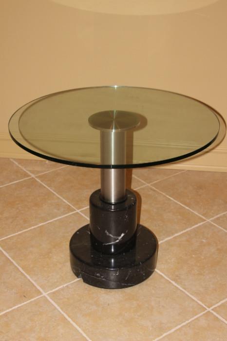 Furniture – Round side table. Glass table top with one pedestaled leg. Piece is metal with a dark marble base, visible chip in marble.