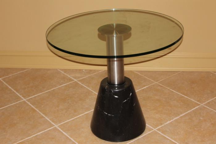 Furniture – Round side table. Glass table top with one pedestaled leg. Piece is metal with a dark marble base, visible chip in marble.
