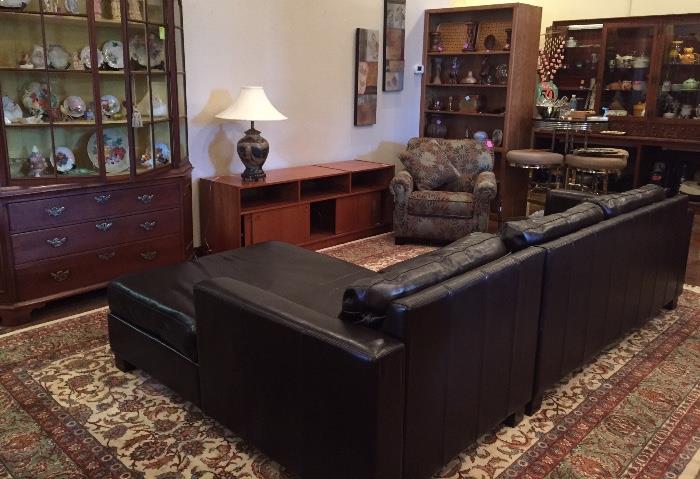 Antique China cabinet filled with bird figurines and plates, dark brown vinyl sofa with chaise, teak media holder, floral chair. 