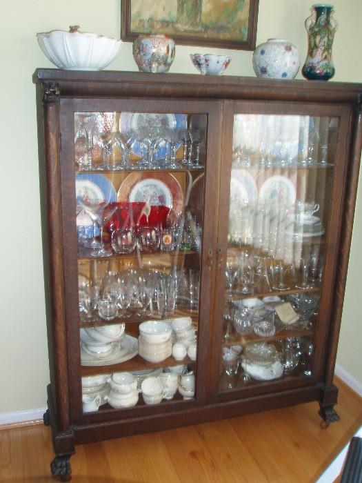 Delightful oak china cabinet with claw feet and lion heads at the top. One door- glass needs replacement- retains original finish- MANY pieces of glassware, stemware and pottery