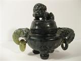 54.	Jade Elephant Incense Pot, circa 1940.               Pre-sale Est. $475
This multi-toned dark green jade incense pot with Elephant handles and temple dog finial has striations that appear to be silver, and others even a gold cast. The feet are round pegs, and the mythological face with a crown surrounds the head. The jade shades from extremely light green (almost translucent) to the very dark with mineral present throughout. 6-3/8” wide and 5-1/4” tall.

