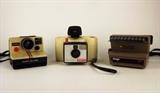 Item Description: A group of three retro Polaroid Land Cameras includes a Swinger model 20 with a wrist strap, a One Step with neck strap and an Amigo 620 with neck strap.
