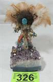 Hand Crafted Kachina on Geode Base
A beautiful Kachina Doll created with a sparkling gold and copper lame costume. The face of the doll is a polished turquoise stone. Appears to be hand carved and decorated. The dolls sits atop a great piece of geode rock with purple crystals. The piece is not signed. Measures approximately 8" tall. The feathers are frayed.
From the internet: "Kachinas are spirits or personifications of things in the real world. A kachina can represent anything in the natural world or cosmos, from a revered ancestor to an element, a location, a quality, a natural phenomenon, or a concept. There are more than 400 different kachinas in Hopi and Pueblo culture. The local pantheon of kachinas varies in each pueblo community; there may be kachinas for the sun, stars, thunderstorms, wind, corn, insects, and many other concepts. Kachinas are understood as having humanlike relationships; they may have uncles, sisters, and grandmothers, and may marry and have children. Although